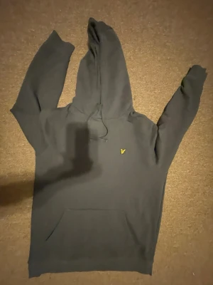 Mörkgrå hoodie från Lyle & Scott - Snygg mörkgrå hoodie från Lyle & Scott med klassisk gul logga på bröstet. Hoodien har dragsko i huvan och en stor magficka framtill. Perfekt för en chill och avslappnad stil. Materialet känns mjukt och skönt mot huden.
