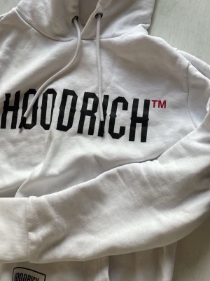 Vit loungewear-set från Hoodrich - Snyggt vitt loungewear-set från Hoodrich med stor svart logga på tröjan och mindre tryck på byxan. Setet består av en hoodie med dragsko och matchande mjukisbyxor med fickor och tryckdetaljer. Perfekt för chill dagar hemma eller streetwear-stil.