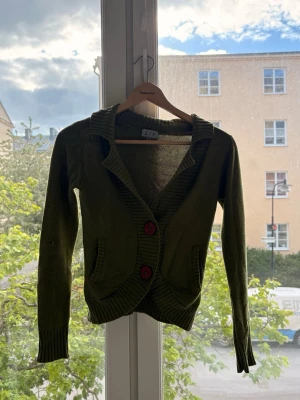 Olivgrön kofta från Z.I.P. XS - Olivgrön stickad kofta från Z.I.P. med två stora röda knappar framtill och ribbade muddar. Koftan har två fickor och en bred krage som ger en cool och avslappnad look. Perfekt att slänga över en t-shirt för extra stil.