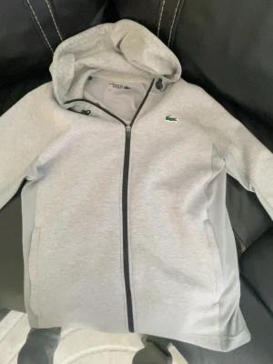 Grå hoodie från Lacoste Sport - Snygg ljusgrå hoodie från Lacoste Sport med dragkedja framtill och klassisk krokodil-logga på bröstet. Hoodien har justerbar huva med snören och sidofickor. Tillverkad i mjukt och bekvämt material, perfekt för en avslappnad stil.