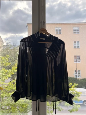 Svart transparent blus från Barro - Snygg svart transparent blus från Barro med dekorativa sömmar på krage och manschetter. Blusen har långa ärmar, knytband framtill och plisserade detaljer upptill. Perfekt för dig som gillar en edgy och trendig stil.