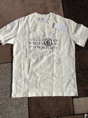 Vit t-shirt Maison Margiela handtryck - Säljer en vit t-shirt från Maison Margiela med ikoniskt handtryck och siffror på bröstet där nummer 6 är markerat. T-shirten har rund hals och är tillverkad i mjuk bomull. Perfekt för dig som gillar unika designer och streetwear vibes. Passar M/L