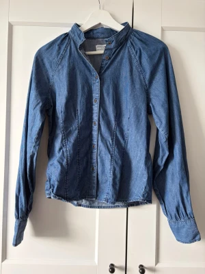 Blå jeansskjorta från Holly & Whyte - Snygg blå jeansskjorta från Holly & Whyte by Lindex. Skjortan har klassisk krage, långa ärmar och knappar framtill. Tillverkad i mjuk bomull som ger en avslappnad men ändå stilren look. Perfekt att matcha med både jeans och kjol.