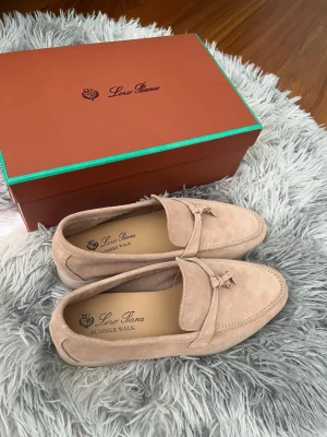Beige loafers från Loro Piana - Snygga beige loafers från Loro Piana i modellen Summer Walk. Skorna är gjorda i mjuk mocka och har en klassisk design med dekorativ detalj framtill. Perfekta för en stilren och avslappnad look.