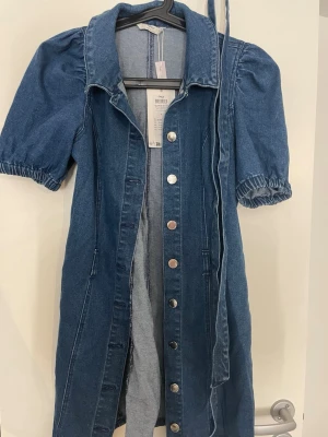 Blå jeansklänning med puffärm - Snygg blå jeansklänning från only med puffärmar och knappar framtill. Klänningen har krage, kort ärm med resår och medföljande bälte i midjan. Perfekt för dig som gillar en avslappnad men trendig stil.