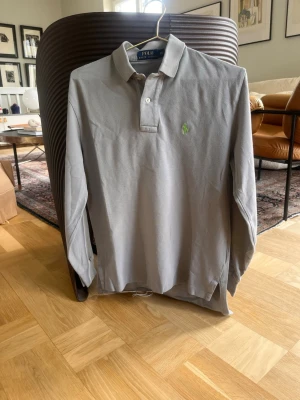 Grå långärmad skjorta från Polo Ralph Lauren - Snygg grå långärmad skjorta från Polo Ralph Lauren med klassisk krage och knappar framtill. Skjortan har en liten grön logga broderad på bröstet och är tillverkad i bomull. Perfekt för en stilren look.