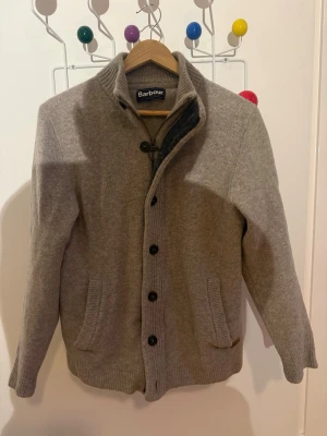 Grå kofta från Barbour med knappar - Snygg grå kofta från Barbour med hög krage och stora svarta knappar. Koftan har två fickor framtill och ribbade muddar vid ärmslut och nederkant. Tillverkad i mjukt material som känns riktigt skönt mot huden. Perfekt för lager på lager och stilren look.