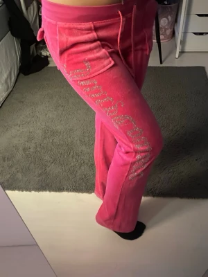 Juicy Couture rosa mjukisbyxor - Säljer ett par ikoniska rosa mjukisbyxor från Juicy Couture i mjuk velour. Byxorna har raka ben, dragsko i midjan och fickor fram. Längs benet står det 'Juicy' i glittriga stenar för extra bling. Perfekta för dig som vill ha en trendig och bekväm look.