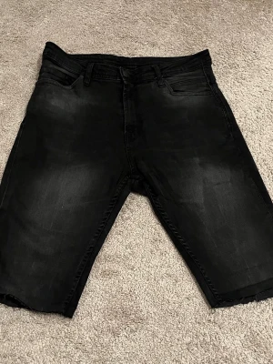 Svarta jeansshorts från Zara - Säljer ett par svarta jeansshorts från Zara  med klassisk femficksdesign och råa benslut. Shortsen har en mörk tvättad look och normal passform. Perfekta för en avslappnad stil under sommaren.