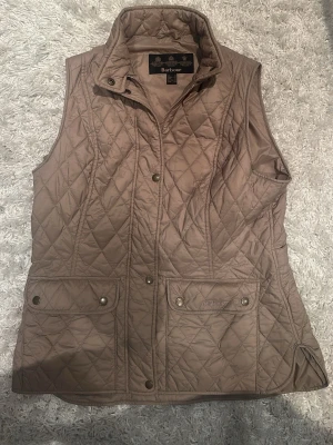 Beige quiltad dunväst från Barbour - Stilren beige dunväst från Barbour med klassiskt quiltat mönster, hög krage och två stora fickor framtill med tryckknappar. Västen har en feminin passform och diskret Barbour-logga på ena fickan. Perfekt lager-på-lager-plagg för kyliga dagar.