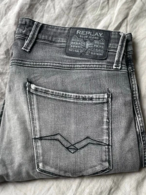 Replay Jeans - Snygga grå jeans från Replay, modell Anbass med slim fit passform. Jeansen har klassisk femficksdesign, detaljerade sömmar på bakfickan och en cool tvättad look. Perfekta för dig som gillar en modern och avslappnad stil.