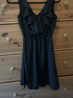 Svart volangklänning med v-ringning - Snygg svart kortklänning med v-ringning och volangdetaljer framtill. Klänningen har ärmlös design och markerad midja för en feminin siluett. Perfekt för dig som gillar stilrena och trendiga plagg.