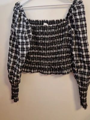Svartvit smockad topp med puffärm - Trendig svartvit rutig topp med smockad kropp och långa puffärmar. Toppen har fyrkantig urringning och elastiska muddar vid ärmsluten. Perfekt för dig som gillar en cool och stilren look. Materialet är lätt och känns skönt mot huden.