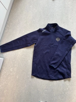 Marinblå träningströja från CCM - Marinblå långärmad träningströja från CCM med logga på bröstet och vit CCM-text på axeln. Tröjan har en praktisk half zip-dragkedja och stårkrage. Perfekt för träning eller uppvärmning.