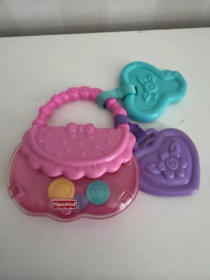 Fisher-Price leksaksväska med hjärtan - Söt leksaksväska i plast från Fisher-Price i rosa, lila och turkos. Väskan har handtag och två hjärtformade hängen med blommönster. Perfekt för små barn som gillar att leka med accessoarer.