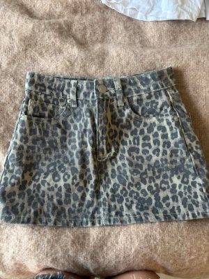 Leopardmönstrad kortkjol i denim - Snygg kortkjol i denim med leopardmönster. Kjolen har klassisk femficksdesign, bälteshällor och knapp framtill. Perfekt för dig som vill sticka ut med en trendig och djurinspirerad look. vet inte storlek men den är ganska liten 