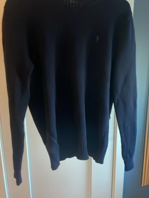 Mörkblå stickad tröja från Polo Ralph Lauren - Klassisk mörkblå stickad tröja från Polo Ralph Lauren med rund halsringning och diskret broderad logga på bröstet. Tröjan har långa ärmar och ribbade muddar vid ärmslut och nederkant. Perfekt för lager-på-lager-stil. PRISET ÄR EJ HUGGET I STEN, KOM MED PRISFÖRSLAG SÅ KAN VI TA DET DÄRIFRÅN!!