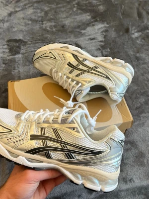 Asics Gel Kayano 14 White/Graphite Grey  - Säljer ett par fräscha Asics sneakers i vitt och silver med svarta detaljer. Skorna har klassisk mesh- och syntetöverdel, snygga kontrastpaneler och tydlig snörning. Perfekta för dig som gillar sportig och trendig stil.