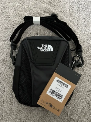 Svart axelväska från The North Face - Snygg svart axelväska från The North Face med justerbar rem och coolt logotryck framtill. Väskan har mesh-detaljer på sidorna och är tillverkad i slitstarkt syntetmaterial. Perfekt för att bära dina viktigaste grejer när du är på språng.