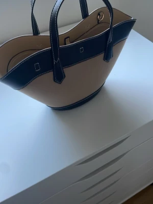 Beige och svart handväska med gulddetaljer - Säljer en snygg beige handväska från H&M med svarta detaljer och kontrastsömmar. Köpte den för 399! Använd 1 gång. Den kommer även med ett långt axelband.