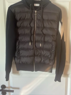Svart cardigan från Moncler - Snygg svart  cardigan från Moncler med vadderad front, stickade ärmar och huva. Dragkedja framtill, två fickor med knapp och coolt vitt zigzag-mönster på ena ärmen samt Moncler-logga. Perfekt för kalla sommar och vinter dagar.