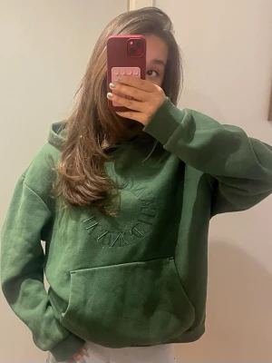 Mörkgrön hoodie - Snygg mörkgrön hoodie med broderad text framtill i samma färg. Hoodien har en stor känguruficka och en klassisk huva. Perfekt för en avslappnad och trendig look. Materialet ser ut att vara mjukt och skönt. Lite nopprig då den e använd men inget farligt därav priset . Ovserize storlek