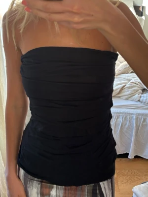 Svart tubtopp från Young Blood - Snygg svart tubtopp från Young Blood med draperad effekt. Toppen är axelbandslös och sitter tight mot kroppen, perfekt för en trendig och stilren look. Mjukt och stretchigt material som formar sig efter kroppen.