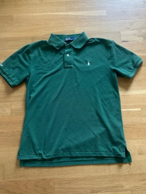 Grön pikétröja från Polo Ralph Lauren - Klassisk grön pikétröja från Polo Ralph Lauren med vit broderad logga på bröstet och 'Polo' broderat på ärmen. Tröjan har krage, två knappar och korta ärmar. Perfekt till en avslappnad men ändå stilren look.