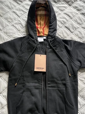 Svart hoodie från Burberry med dragkedja - Svart hoodie från Burberry med klassiskt rutigt foder i huvan och dragkedja framtill. Stora fickor, justerbar huva med snören och diskret Burberry-logga på bröstet. Perfekt för en stilren och avslappnad look. Tveka inte med att ställa frågor och kom med bud!