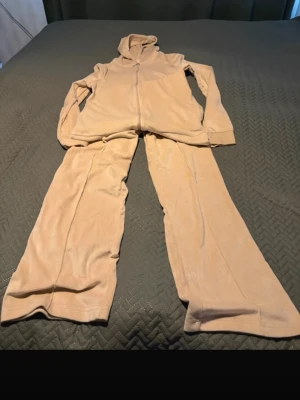 Beige mjukis-set med hoodie och byxor - Mysigt loungewear-set i beige med zip-hoodie och matchande vida byxor. Setet är tillverkat i ett mjukt och skönt material, perfekt för chill dagar hemma. Hoodien har dragkedja och långa ärmar, byxorna har avslappnad passform.