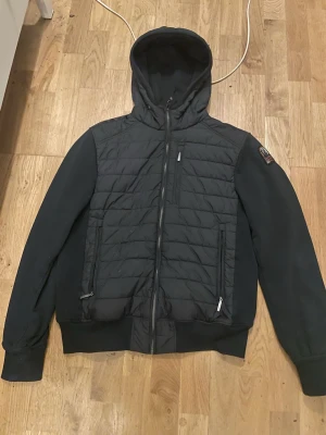 Parajumpers cardigan  - Snygg svart cardigan från Parajumpers med huva och dragkedja framtill. Jackan har quiltad front, tre dragkedjefickor och ribbade muddar. Klassisk Parajumpers-logga på ärmen. Perfekt för alla dagar.  Med en qr kod som tar dig till deras hemsida för bevis att den är äkta 