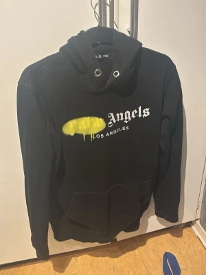 Palm Angels svart hoodie med tryck - Svart hoodie från Palm Angels med stor huva och metallöljetter. Framsidan har ett vitt Los Angeles-tryck och en gul spray paint-detalj. Klassisk känguruficka framtill och avslappnad passform. Perfekt för en streetwear-look.