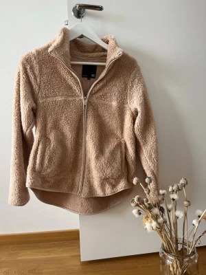 Beige teddyjacka från 157 - Mysig beige teddyjacka från 157 med dragkedja framtill och hög krage. Jackan har två fickor och är tillverkad i fluffigt teddy-material som ger en avslappnad och trendig look. Perfekt för lager-på-lager-stil under kyliga dagar.