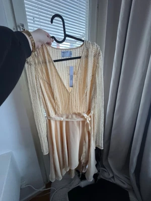 Champagnefärgad klänning med pärlor - Superfin champagnefärgad kortklänning från ASOS med långa ärmar, v-ringning och pärldetaljer i ränder. Klänningen har ett satinband i midjan som knyts och ger en snygg siluett. Perfekt för dig som vill sticka ut med elegans. Köpt för 999kr