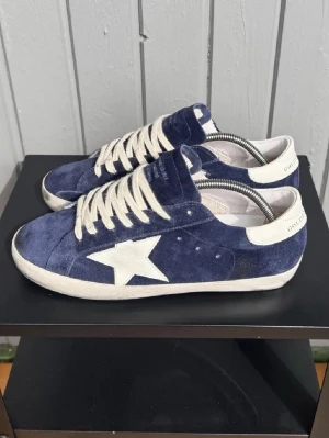 Blå Golden Goose sneakers - Snygga blå sneakers från Golden Goose med vit stjärndetalj på sidan och vita skosnören. Skorna är i mocka med vita detaljer på häl och sula. Sjukt populära och eftertraktade - 9/10-storlek 42 - inget og medföljs!!