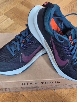 Nike Juniper Trail 2  svart/lila - Nike Juniper Trail 2 Next Nature är en bekväm terränglöparsko med en blandning av meshmaterial, syntetiskt läder och textilmaterial i överdelen, vilket garanterar utmärkt slitstyrka och andningsförmåga under löprundan. Yttersulan är tillverkad av gummimaterial som ger bra fäste och grepp i tuff terräng. Storlek EUR 39 cm 25 