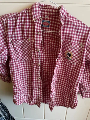 Rutig skjorta från Grants, röd/vit - Röd och vit rutig skjorta från Grants med klassisk krage, två bröstfickor och knappar framtill. Skjortan har lång ärm och ett litet broderat märke på ena fickan. Perfekt för en avslappnad och cool stil.