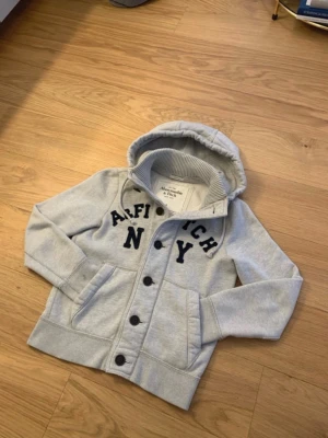 Grå hoodie från Abercrombie & Fitch - Snygg ljusgrå hoodie från Abercrombie & Fitch med stora mörkblå bokstäver på bröstet. Tröjan har knappar framtill, två fickor och en mysig huva med dragsko. Tillverkad i mjukt sweatshirtmaterial, perfekt för chill dagar.