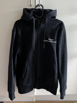 Svart Peak Performance zip-tröja - Svart tröja från Peak Performance med dragkedja. Vit text på bröstet och liten logga på ärmen. Skön och stilren.