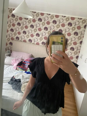 Svart omlottopp med volangärm - Svart omlottopp med v-ringning och volangärmar. Toppen har knyt i nacken och är tillverkad i ett lätt, luftigt material som ger en avslappnad och trendig look. Perfekt att styla med jeans eller kjol för en snygg outfit.