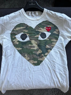Cdg camouflage T-shirt - Vit t-shirt från Comme des Garçons Play med stort hjärta i camouflage-mönster och ögon på bröstet. Liten röd hjärtdetalj med ögon ovanför. Klassisk rund hals och korta ärmar. Bara att höra av er vid frågor, inte används på 1 år.