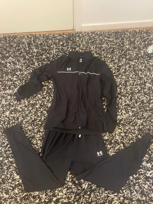 Svart träningsset från Under Armour - Säljer ett svart träningsset från Under Armour med jacka och byxor. Jackan har dragkedja, ståkrage och en vit detalj över bröstet. Byxorna har resår i midjan och logga på benet. Perfekt för träning eller uppvärmning.