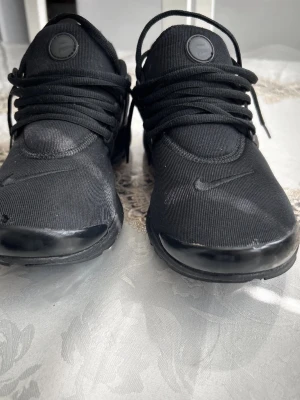 Svarta Nike Presto sneakers - Svarta Nike Presto sneakers med mesh-ovandel och klassisk snörning. Skorna har en diskret Nike-logga på sidan och framtill samt en flexibel sula för extra komfort. Snygg minimalistisk design som passar till streetwear och casual outfits.