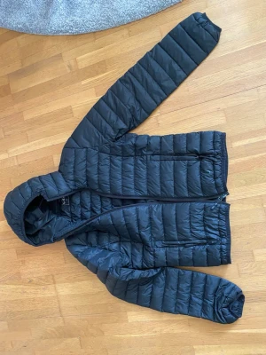 Svart dunjacka med huva - Säljer en svart tunn dunjacka med huva och dragkedja framtill. Jackan har två sidofickor med dragkedja och quiltad design. 