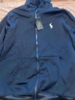 Mörkblå Polo hoodie  - Snygg mörkblå hoodie från Polo Ralph Lauren med dragkedja framtill och klassisk vit logga på bröstet. Hoodien har huva med dragsko och känguruficka. Perfekt för en avslappnad och stilren look.