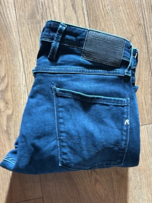 Blå slim fit jeans från Replay - Snygga blå slim fit jeans från Replay med klassisk femficksdesign och diskret logga på bakfickan. Jeansen har en mörk tvätt med lätt slitningar framtill och är tillverkade i mjukt denimtyg. Perfekta för en trendig och avslappnad stil. W32 L32
