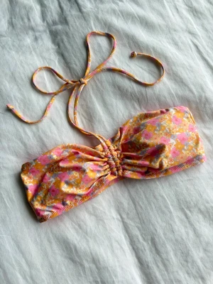 Blommig bandeau bikiniöverdel rosa/gul - Söt bandeau bikiniöverdel med blommigt mönster i rosa, gult och orange. Toppen har snörning i nacken och rynkade detaljer framtill för en snygg passform. Perfekt för sommarens strandhäng!