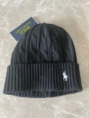 Svart kabelstickad mössa Polo Ralph Lauren - Snygg svart kabelstickad mössa från Polo Ralph Lauren med uppvikt kant och klassisk broderad logga i vitt framtill. Tillverkad i mjukt material som håller dig varm och stilren under kalla dagar.