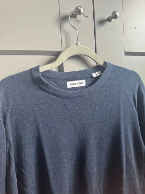 Mörkblå tröja från Jack & Jones - En stilren mörkblå tröja från Jack & Jones med rund halsringning och långa ärmar. Tröjan har ribbade muddar vid ärmslut och nederkant, vilket ger en snygg passform. Perfekt att matcha med jeans eller chinos för en clean look.