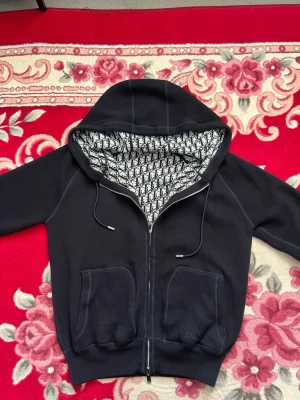 Svart hoodie från Dior med monogram - Snygg svart hoodie från Dior med dragkedja och två stora fickor framtill. Insidan av huvan har ett ikoniskt svartvitt Dior-monogram som ger en exklusiv vibe. Tillverkad i mjukt material och har justerbara snören i huvan. Perfekt för dig som gillar streetwear och designerplagg. Äkta hoodie 
